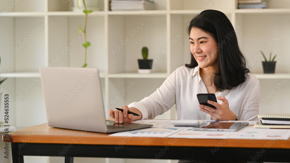 Pretty asian female employee holding mobile phone and using laptop on wooden office desk