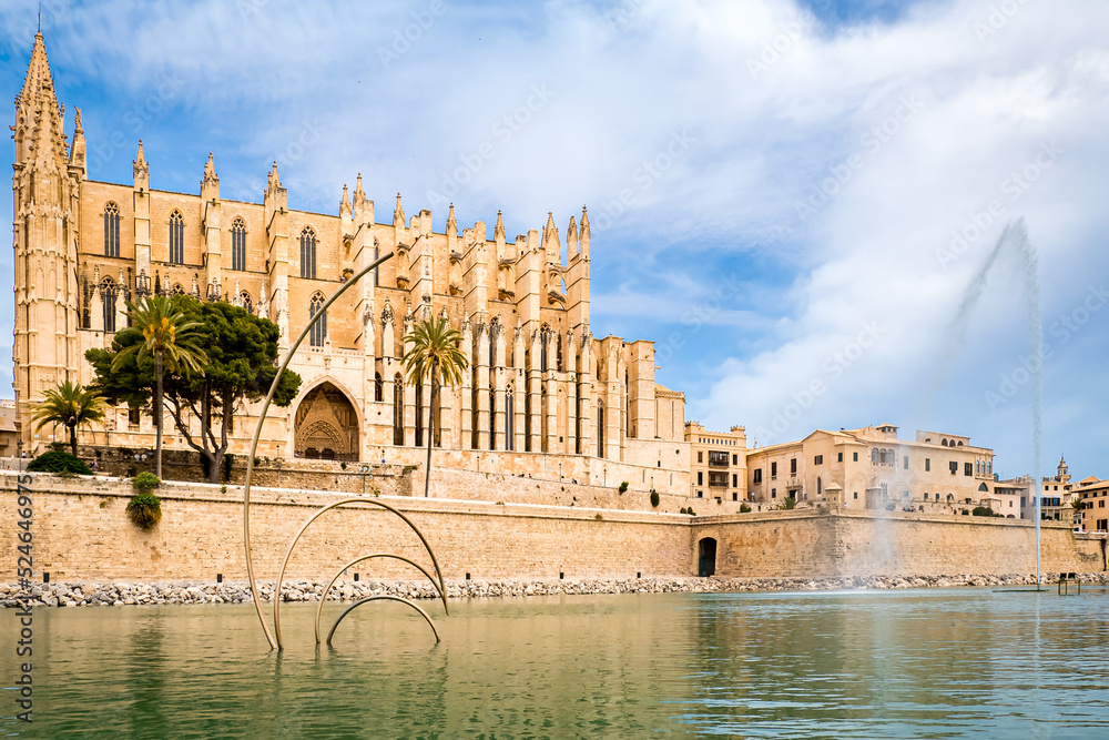 Panoramic view over the pond with art and a fountain of the Parc de la Mar in the city of Palma de Mallorca with the gothic style cathedral La Seu de Mallorca in front of cloudy sky in the background.