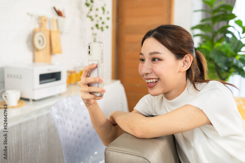 Happy and cheerful woman is using a phone sitting on a sofa.Asian young woman looking mobile phone for shopping online at home. Happy and cheerful woman is using a phone sitting on a sofa.Asian young woman looking mobile phone for shopping online at home.