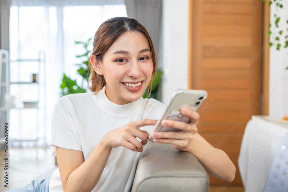 Happy and cheerful woman is using a phone sitting on a sofa.Asian young woman looking mobile phone for shopping online at home. Happy and cheerful woman is using a phone sitting on a sofa.Asian young woman looking mobile phone for shopping online at home.