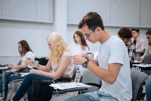 A young guy with glasses during a physics test takes pictures of a test on his smartphone, to show it to his friends from other college groups