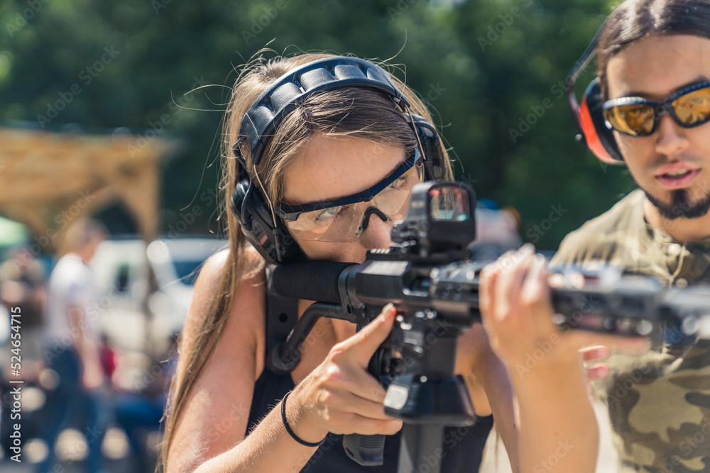 Obraz premium Male instructor observing woman in safety gear using submachine gun. Target practise. Firearms training at outdoor shooting range. Horizontal shot. High quality photo