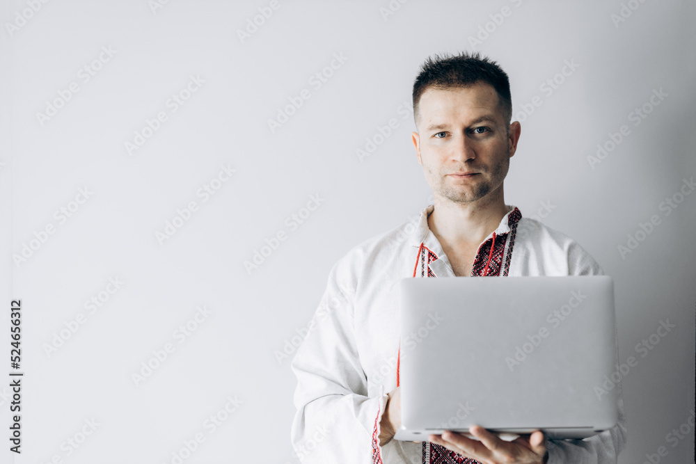 Guy in Ukrainian vyshyvanka stands holding laptop.