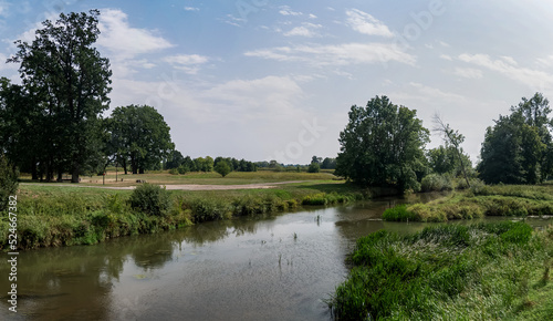 Panorama rzeki Osobłogi w obszarach polnych, w tle zarośla i nieliczne drzewa w porze letniej na tle nieznacznie pochmurnego nieba