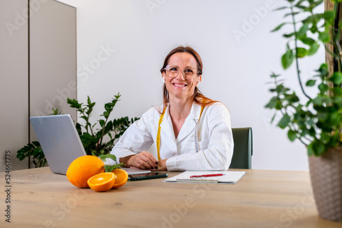 Portrait of smiling young female nutritionist in her office in medical gown with fresh oranges. Dietologist making a diet plan