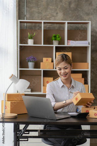 New business owner Asian woman working at home office Packaging on background SME entrepreneur online shopping or freelance work concept Young woman happy after new order from customer