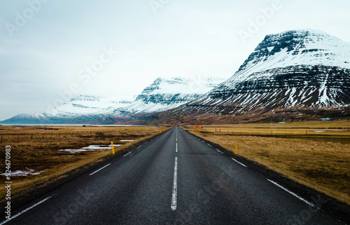 Panoramic winter photo of road leading along coast of lake to volcanic mountains. High rocky peaks covered with snow layer mirroring on water surface. Driver's point of view on Ring road, Iceland.
