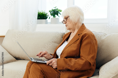 an attentive elderly woman with glasses is mastering new technologies using a laptop while sitting on the couch at home in a comfortable environment in a brown suit