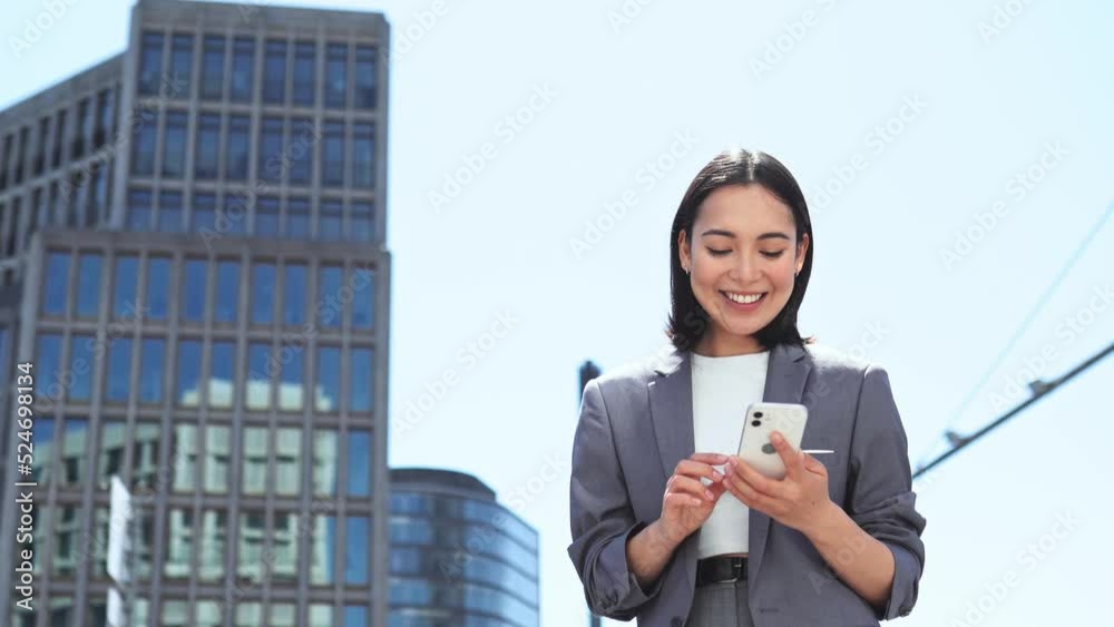 Young happy Asian woman professional executive using cell phone receiving good news in mobile message, smiling ethnic businesswoman holding smartphone looking at telephone standing on big city street.