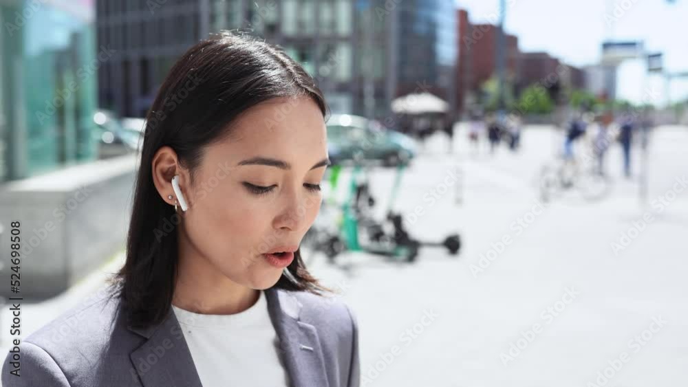 Young professional Asian business woman wearing suit using wireless earbuds on mobile internet data using smartphone talking on mobile phone having chat standing on city street. Mobility