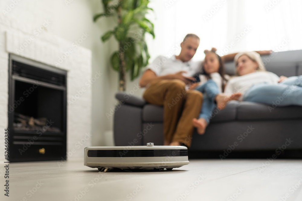 Young family resting on the couch while robotic vacuum cleaner doing its work at home. Household robots concept.