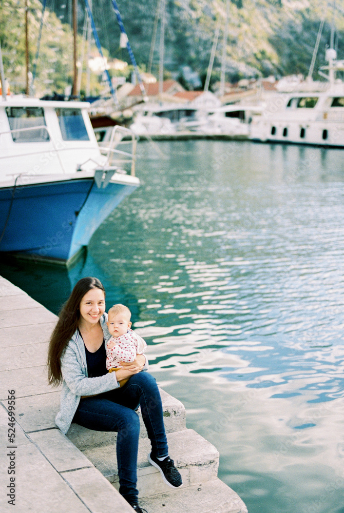 Smiling mother with a baby on her lap sits on the pier near the boat