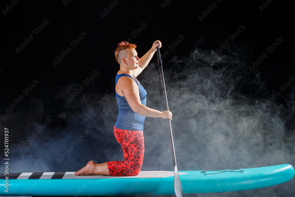 A woman with a mohawk on a sup board on her knees with an oar in the fog on a black background.