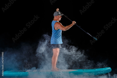 A man in shorts on a sub board with a paddle on a black background in the fog.
