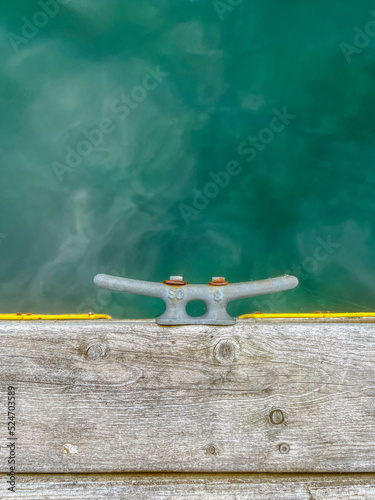 Looking down over a single metal cleat affixed to the side of a wooden pier. Crystal clear turquoise waters are seen below. Yellow protective material lines the side of the dock for protection.
