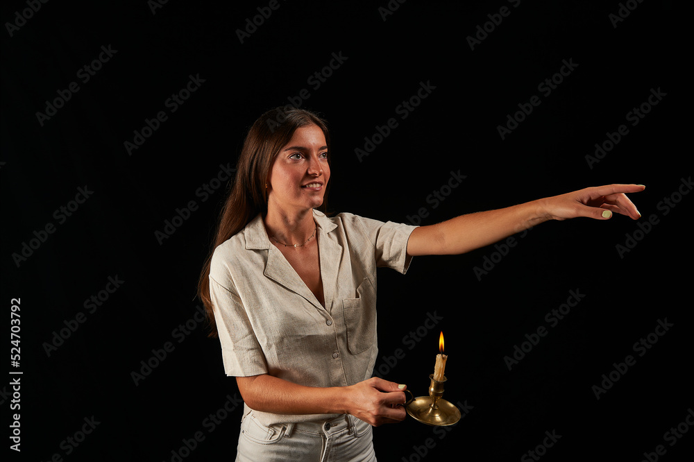 Fototapeta premium young woman lit by an old brass lamp with a candle on a black background points with her left hand
