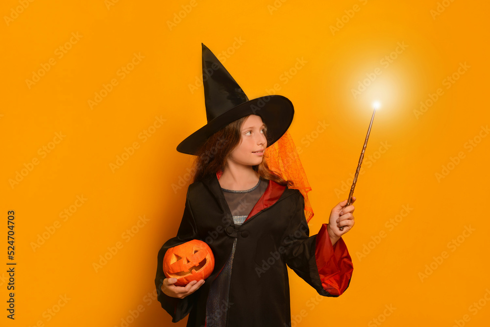 Cute pre-teen girl in wizard student costume holding luminous magic wand or glowing magic stick with carved Halloween pumpkin or Jack o lantern pumpkin and looking at light on yellow background.
