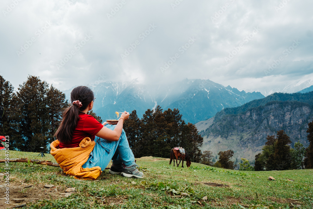 Indian female tourist eating breakfast while enjoying view of Himalayan mountains during trekking. Girl eating noodles, sitting on grass after hiking in mountains at Manali, India 