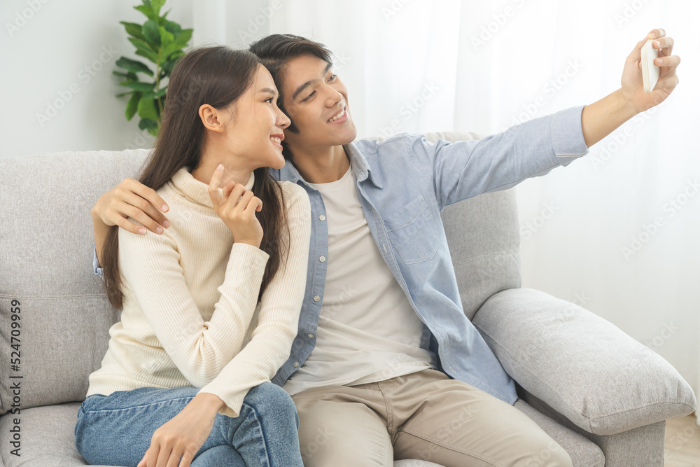 Happy couple love at home, two asian young spending time, bonding to each other romantic on sofa in living room while man embrace woman using smartphone, mobile phone to selfie take a photo together. Happy couple love at home, two asian young spending time, bonding to each other romantic on sofa in living room while man embrace woman using smartphone, mobile phone to selfie take a photo together.