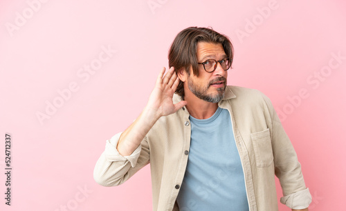 Senior dutch man isolated on pink background listening to something by putting hand on the ear