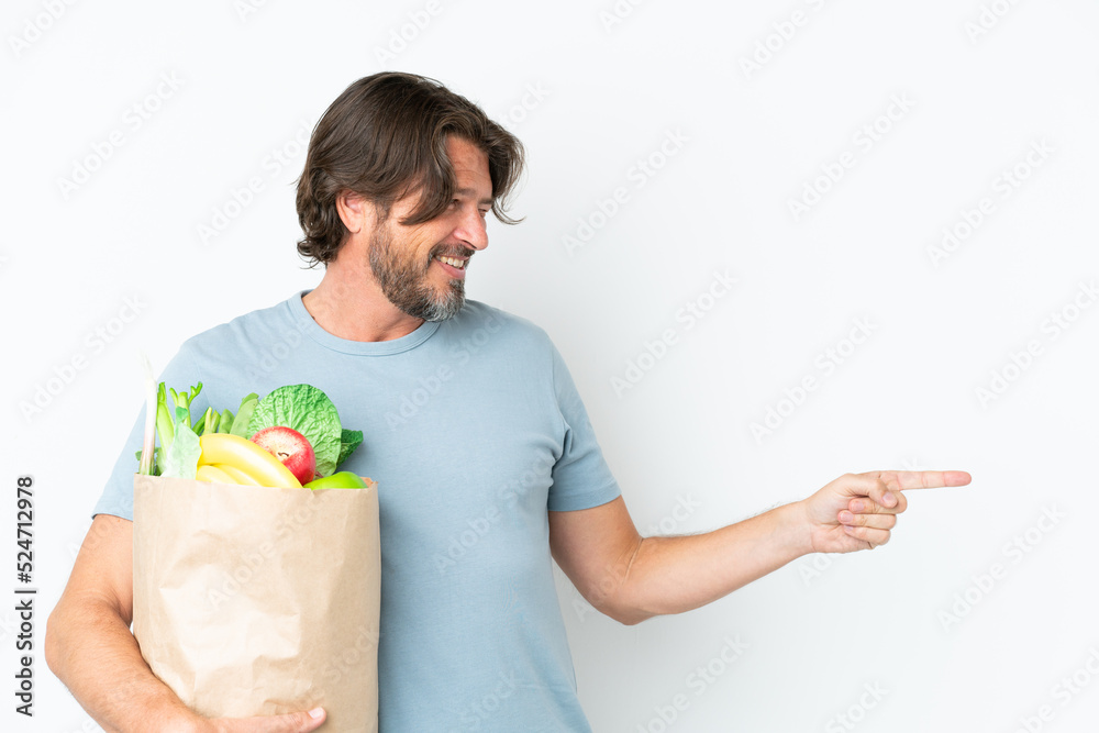 Senior dutch man holding grocery shopping bag over isolated background pointing finger to the side and presenting a product Senior dutch man holding grocery shopping bag over isolated background pointing finger to the side and presenting a product