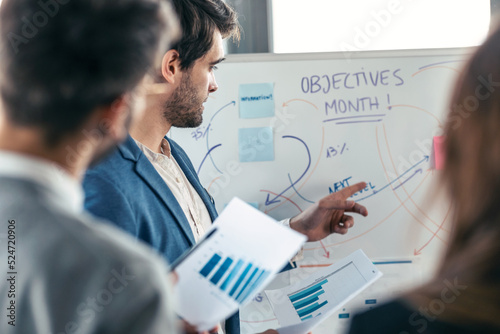 Handsome businessman pointing at white blackboard and explain a project to her colleagues on coworking place.