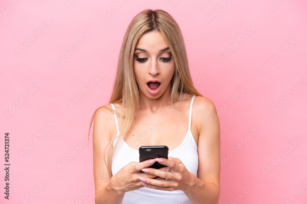 Pretty blonde woman isolated on pink background looking at the camera while using the mobile with surprised expression Pretty blonde woman isolated on pink background looking at the camera while using the mobile with surprised expression