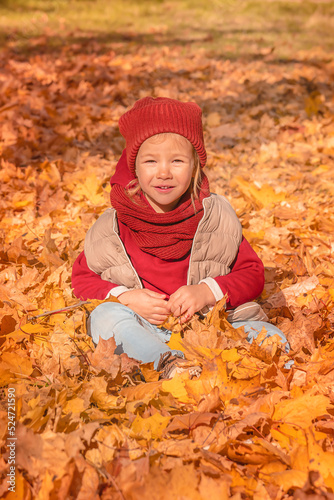 Beautiful child sits on a glade with maple leaves in an autumn park. Happy child girl having fun in autumn park