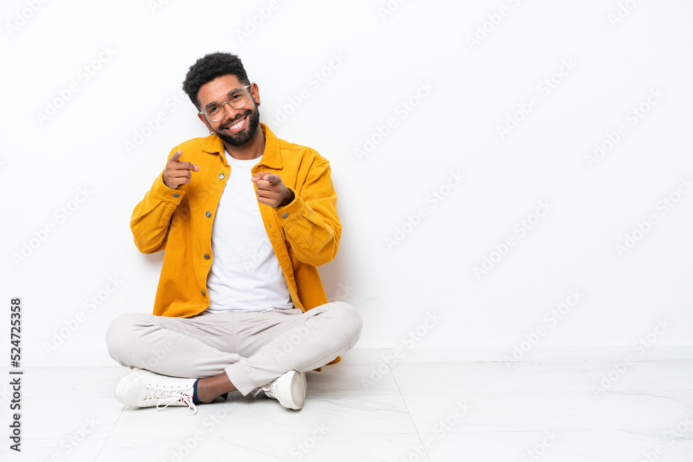 Young Brazilian man sitting on the floor isolated on white background pointing to the front and smiling Young Brazilian man sitting on the floor isolated on white background pointing to the front and smiling