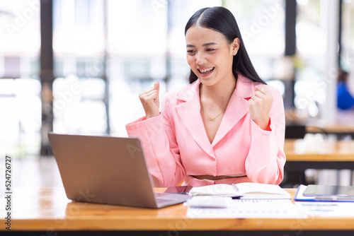Free Download Happy excited successful Asian businesswoman triumphing with a laptop computer smartphone in the workplace office
 - GFXLoad