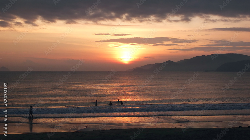  Seacoast before sunrise with silhouettes of swimming and walking  peoples and mountains in the distance .Summer vacation near the sea, traveling concept