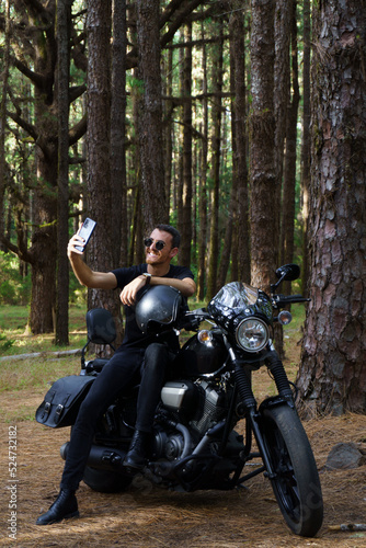 Motero con moto custom haciéndose un selfie en el bosque