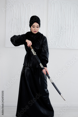 Woman in hijab, black headscarf and dress. The woman is holding a sword in her hand. Stylish young beautiful Muslim woman.