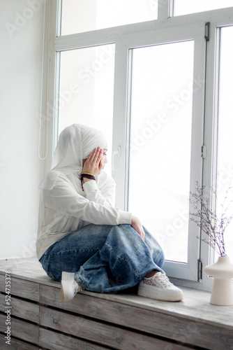 A Muslim woman in a white hijab and white blouse sits on the windowsill and looks out the window. Thoughtfulness, contemplation. © Sarbinaz Mustafina