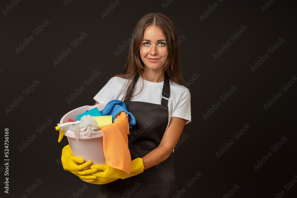 A young woman with long hair in gloves for cleaning, in an apron, with a bucket in her hands. Cleaning products in a bucket. Studio shot. A young woman with long hair in gloves for cleaning, in an apron, with a bucket in her hands. Cleaning products in a bucket. Studio shot.