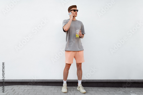 Portrait of handsome confident stylish hipster man in sunglasses talking on the phone, standing guy near the concrete wall. Calm relaxing moment, summer vacation concept