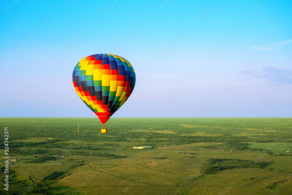 Obraz premium A colorful air balloon is flying in free flight over the field. Bird's-eye view. Multi colored balloon in the blue sky