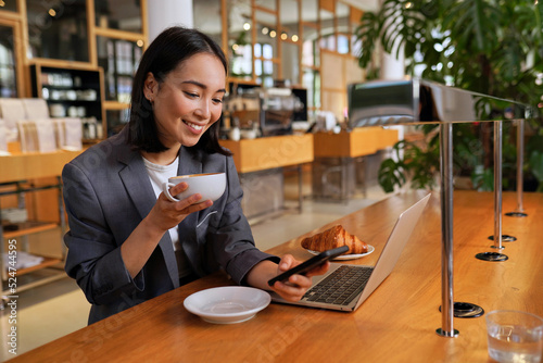 Fotografie Young Asian business woman wearing suit drinking coffee using smartphone in cafe