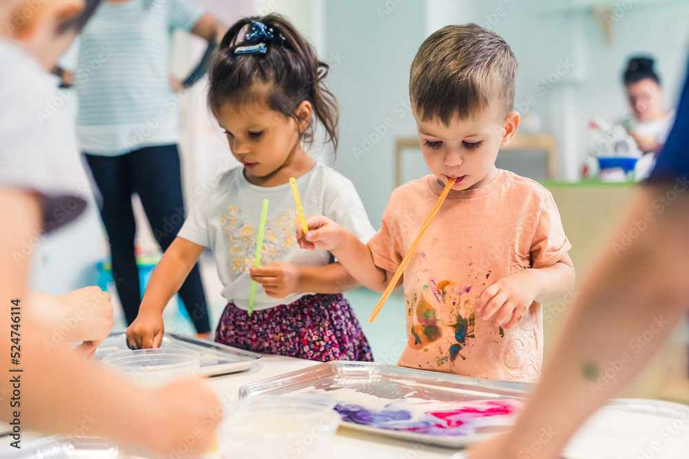 Multi-ethnic group of toddlers milk painting with the teacher helping them, using food coloring for colors. Children finger painting at the nursery school class. High quality photo - obrazy, fototapety, plakaty 