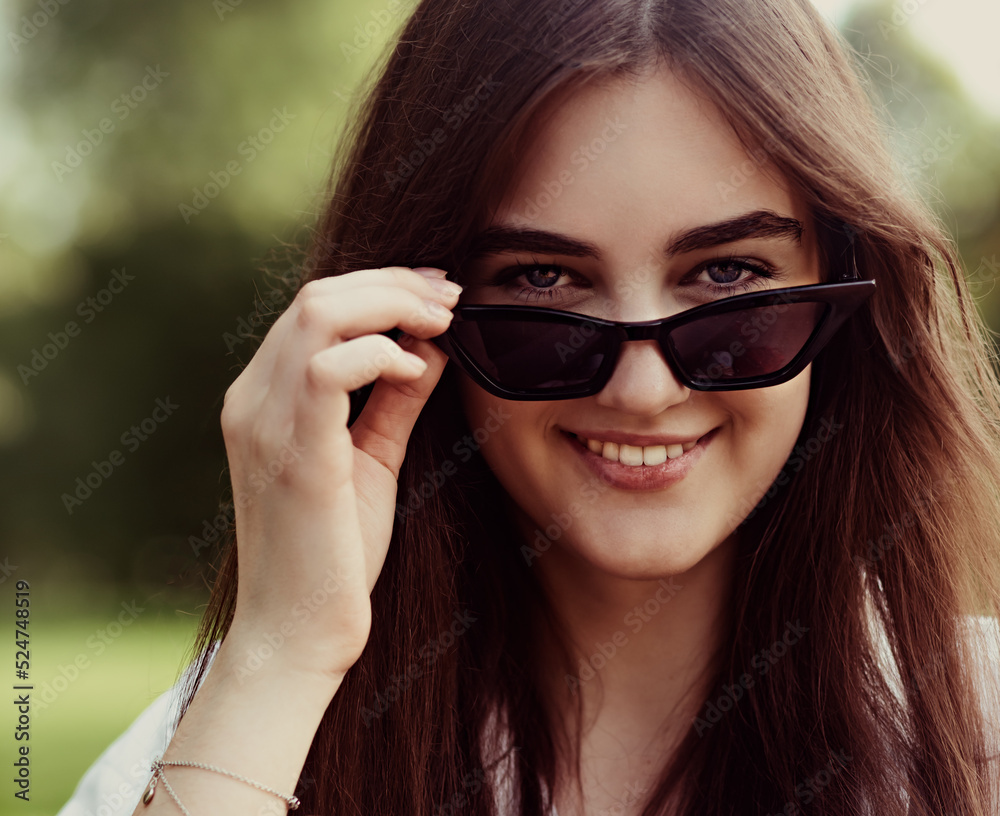Beautiful smiling teen woman looking happy in fashion black sun glasses on green glass outdoors summer trees background in part. Closeup portrait Beautiful smiling teen woman looking happy in fashion black sun glasses on green glass outdoors summer trees background in part. Closeup portrait