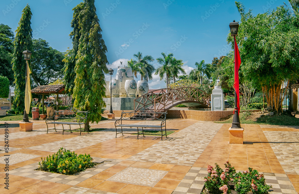 Fototapeta premium A nice and relaxed park with a wooden bridge over a water fountain, Traditional park of Nagarote, Nicaragua. View of a calm park with a small wooden bridge on a sunny day. Nagarote central park