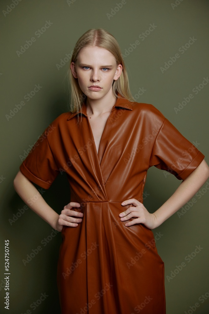 Self-confidence blonde model with blue eyes and evening makeup posing in trendy leather fashion brown palette clothes holding hands on waist looks at camera staying isolated on green background Self-confidence blonde model with blue eyes and evening makeup posing in trendy leather fashion brown palette clothes holding hands on waist looks at camera staying isolated on green background