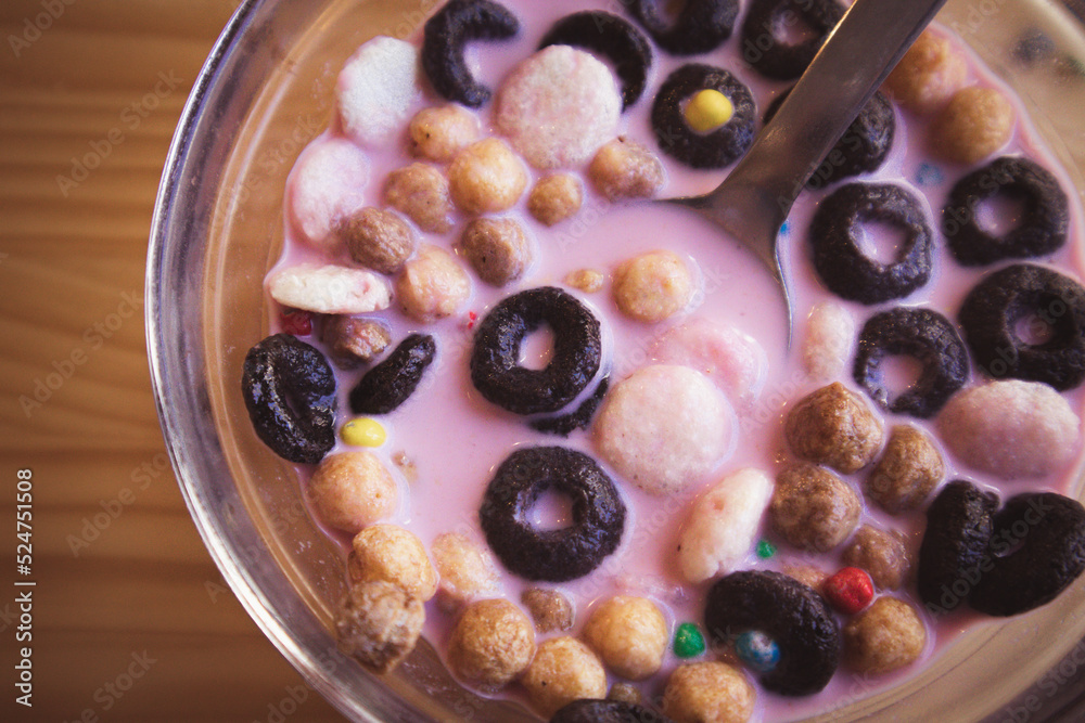 Dry breakfast in glass round plate bowl on a wooden table in cafe. Delicious cornflakes drenched in pink strawberry milk. Healthy breakfast. Food for kids. Different type cereals, multicolored balls. Dry breakfast in glass round plate bowl on a wooden table in cafe. Delicious cornflakes drenched in pink strawberry milk. Healthy breakfast. Food for kids. Different type cereals, multicolored balls.
