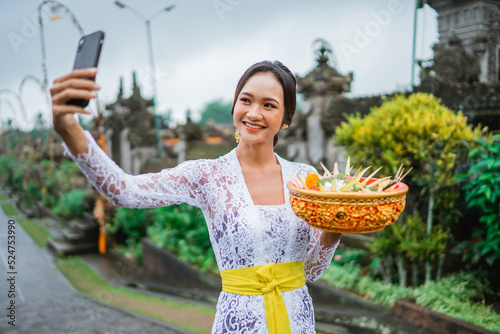 attractive balinese young woman taking her selfie photo with mobile phone while wearing traditional kebaya clothes in bali photo