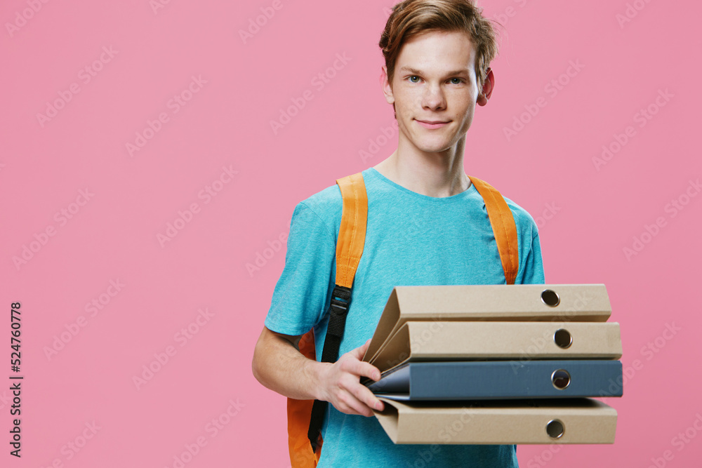 Obraz premium a handsome guy with an orange backpack on his back holds empty note folders in his hands, smiling pleasantly at the camera. Horizontal photo on a pink background with empty space for advertising text