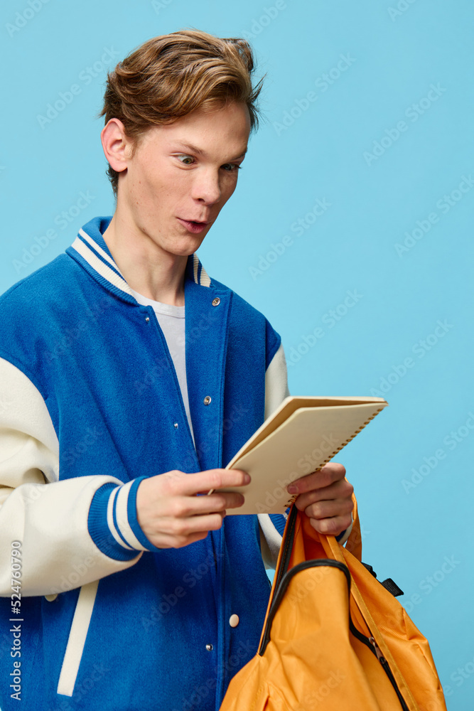 funny student in a bomber jacket stands with a notepad in his hand on a blue background and makes a funny face in an attempt to make out the written text. Training, preparation for the training season funny student in a bomber jacket stands with a notepad in his hand on a blue background and makes a funny face in an attempt to make out the written text. Training, preparation for the training season