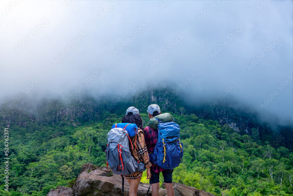 Lover trekker tourist backpack ,hat with trekking stick stand on mountain stone. they adventure trekking  beautiful floating white foggy green forest national park in vacation is eco tourism concept