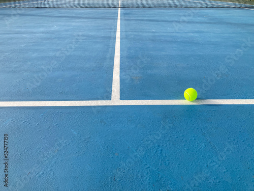 the green tennis ball is on the white line and the black net is in the middle of the blue tennis court in super bright sunlight