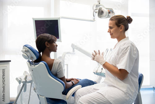 Smiling female patient having consultation at dentist office. Young Caucasian dentist wearing gloves talking to African American woman in dental clinic. Visiting dentist concept
