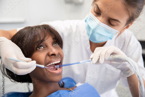 Female dental hygienist using dental hook and suction during cleaning teeth procedure. Young African American woman visiting stomatology clinic. Oral hygiene and dental care concept photo